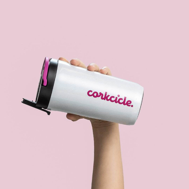 Sparkle Unicorn Magic Cup is a white silicone cup with "Corkcicle" written vertically in pink. The cap part is black and the handle is pink. On the cap part, the drinking straw is raised and the cup is held horizontally in one hand, on a pale pink backgro
